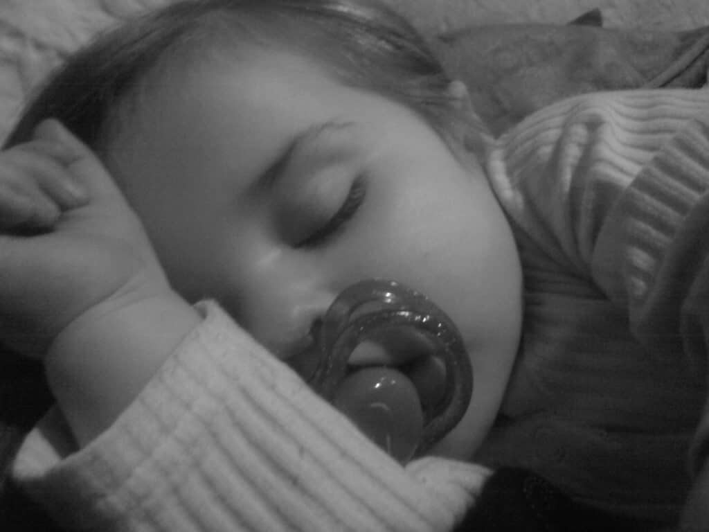A close-up, black and white photo of a young child peacefully sleeping with a dummy in their mouth. The child’s arm is bent and resting near their face, and they are wearing a long-sleeved, ribbed shirt. The image conveys calm and innocence.