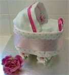 A decorative item shaped like a pram, made with white and pink fabric, lace trim, and a pink ribbon, sits on a silver surface. A pink artificial rose is placed beside it. The background is a tiled wall.