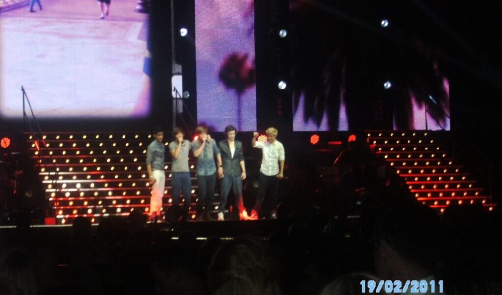 Five young men stand singing on a brightly lit concert stage with red and yellow stair lights. A large screen behind them shows a cityscape and palm trees. Audience heads are visible in the dark foreground. The date “19/02/2011” appears in blue text.