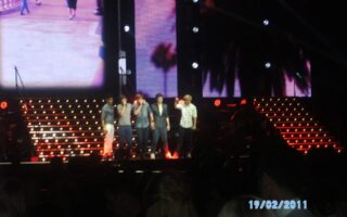 Five young men stand singing on a brightly lit concert stage with red and yellow stair lights. A large screen behind them shows a cityscape and palm trees. Audience heads are visible in the dark foreground. The date “19/02/2011” appears in blue text.