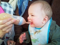 A baby wearing a blue bib and green jacket sits on a brown sofa, being fed brownish food from a bottle with a spoon attachment by an adult. The baby has food on the bib and is leaning forward with its mouth open, ready to eat.