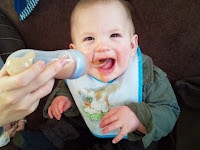A smiling baby wearing a white bib with a blue border and animal design is being fed with a bottle by an adult's hand. The baby has food smeared on their face and bib, and sits on a dark-coloured surface, appearing happy and engaged.