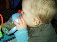 A baby with fair hair, dressed in a blue and grey outfit, is holding and drinking from a blue beaker. The baby is seen from the side, with focus on their head and hands. The background is slightly blurred and dimly lit.