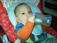 A baby with fair skin and short hair sits in a red and white patterned highchair, wearing a blue bib and striped shirt, holding and drinking from a baby bottle with both hands. The background includes part of a kitchen appliance.