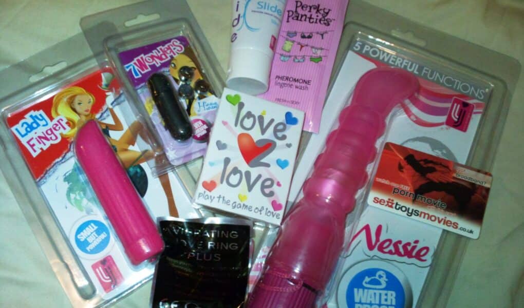 A selection of adult toys and products, including vibrators, lubricant, love-themed playing cards, pheromone cream, a vibrating ring, and a voucher for a sex toy/film website, all arranged on a flat surface with colourful packaging.