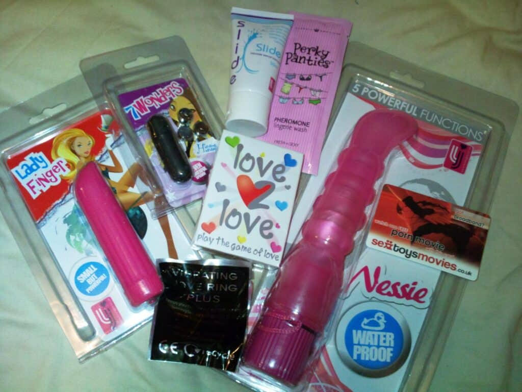 A selection of adult toys and products, including vibrators, lubricant, love-themed playing cards, pheromone cream, a vibrating ring, and a voucher for a sex toy/film website, all arranged on a flat surface with colourful packaging.