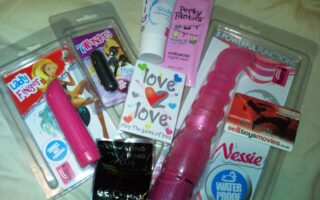 A selection of adult toys and products, including vibrators, lubricant, love-themed playing cards, pheromone cream, a vibrating ring, and a voucher for a sex toy/film website, all arranged on a flat surface with colourful packaging.