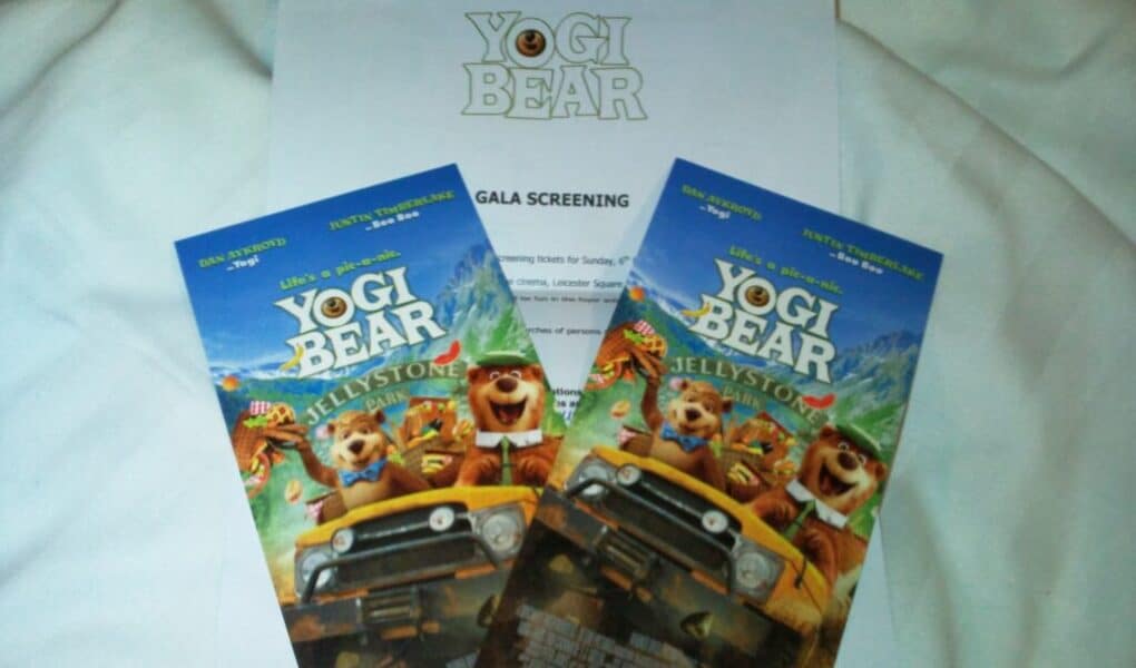 Two colourful “Yogi Bear” cinema tickets featuring animated bears in a car are placed on a white paper labelled “Yogi Bear Gala Screening.” All items rest on a white fabric background. The tickets display the film title and images of Yogi and Boo Boo.