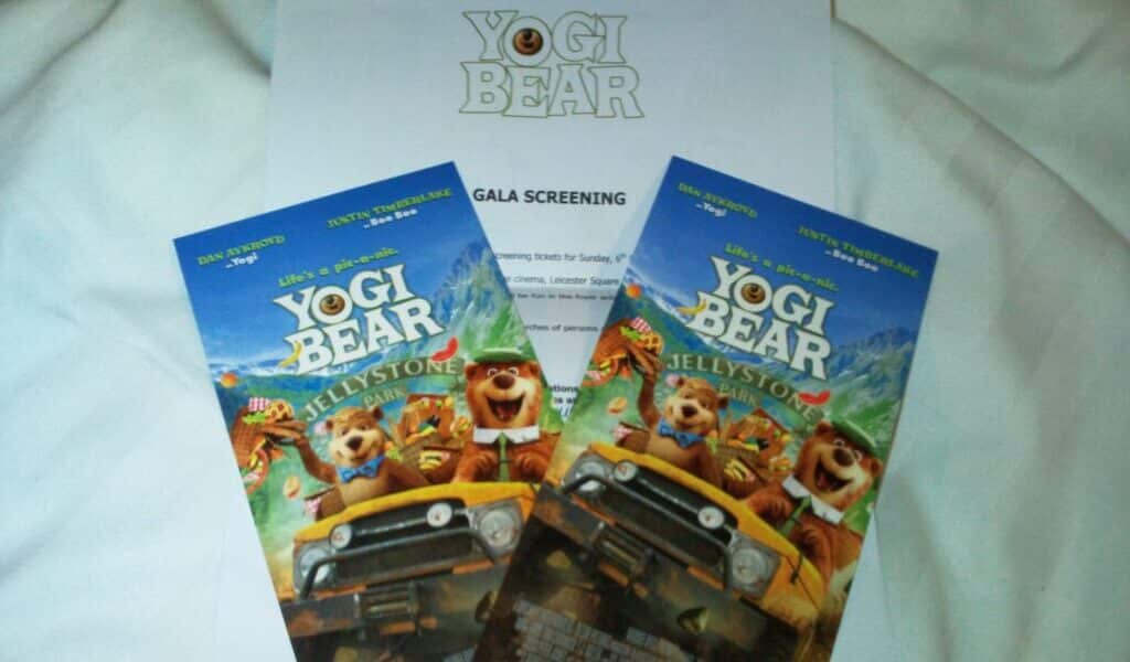 Two colourful “Yogi Bear” cinema tickets featuring animated bears in a car are placed on a white paper labelled “Yogi Bear Gala Screening.” All items rest on a white fabric background. The tickets display the film title and images of Yogi and Boo Boo.