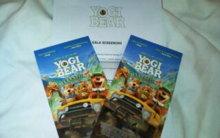 Two colourful “Yogi Bear” cinema tickets featuring animated bears in a car are placed on a white paper labelled “Yogi Bear Gala Screening.” All items rest on a white fabric background. The tickets display the film title and images of Yogi and Boo Boo.