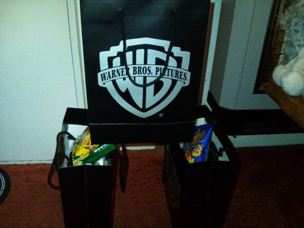 A black Warner Bros. Pictures gift bag and two similar black bags sit on a red carpet. The bags contain visible snacks, including crisps and sweets. Part of a soft toy and a wooden table are visible on the right side of the image.