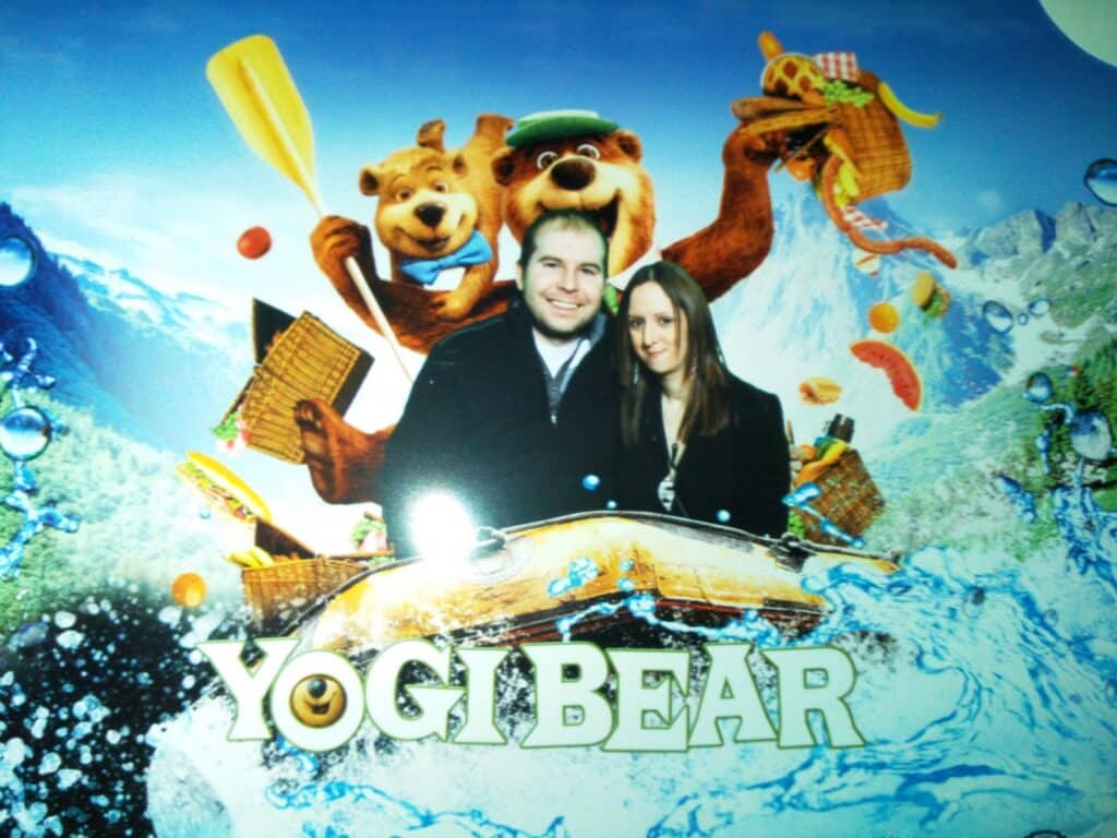 A man and woman pose in front of a “Yogi Bear” display, appearing to ride a boat with cartoon characters Yogi and Boo Boo behind them, holding paddles and picnic items. The background features mountains, water splashes, and the “YOGI BEAR” film title.