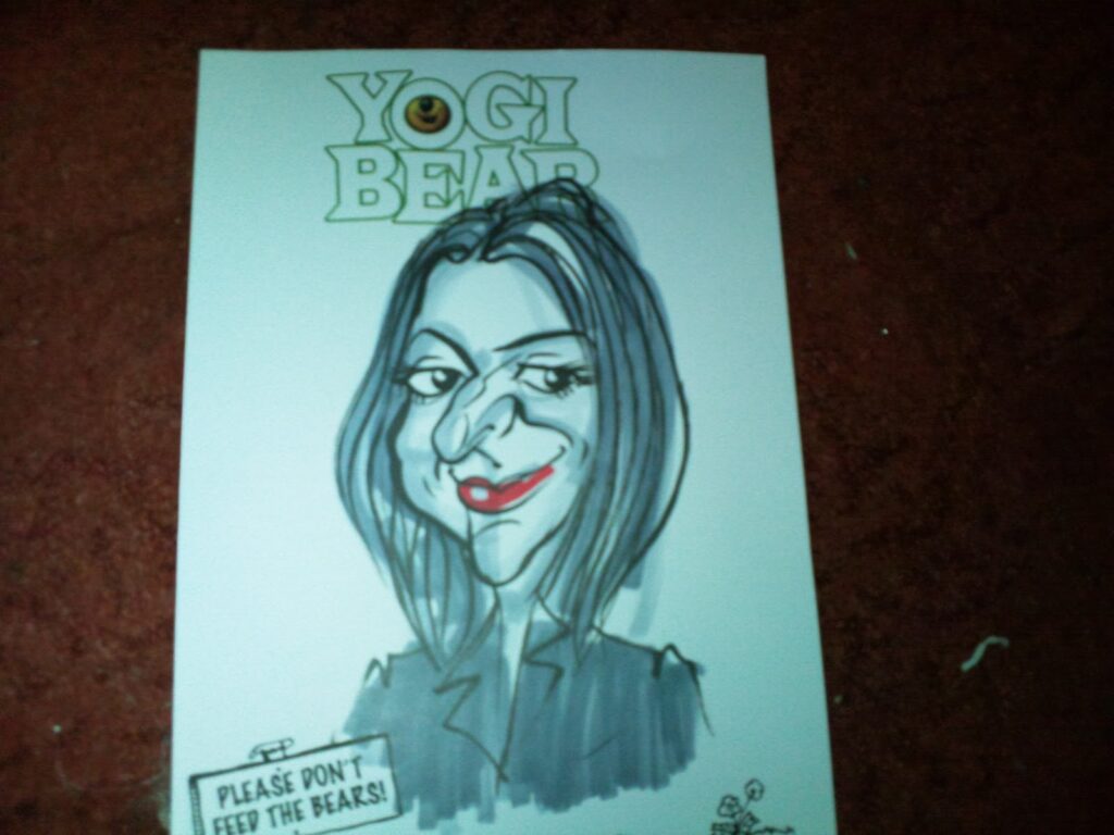 A caricature drawing of a woman with long dark hair, exaggerated facial features, and red lips, wearing a dark outfit. Above her, “YOGI BEAR” is written. A small sign at the bottom left reads, “PLEASE DON’T FEED THE BEARS!” The background is brown.