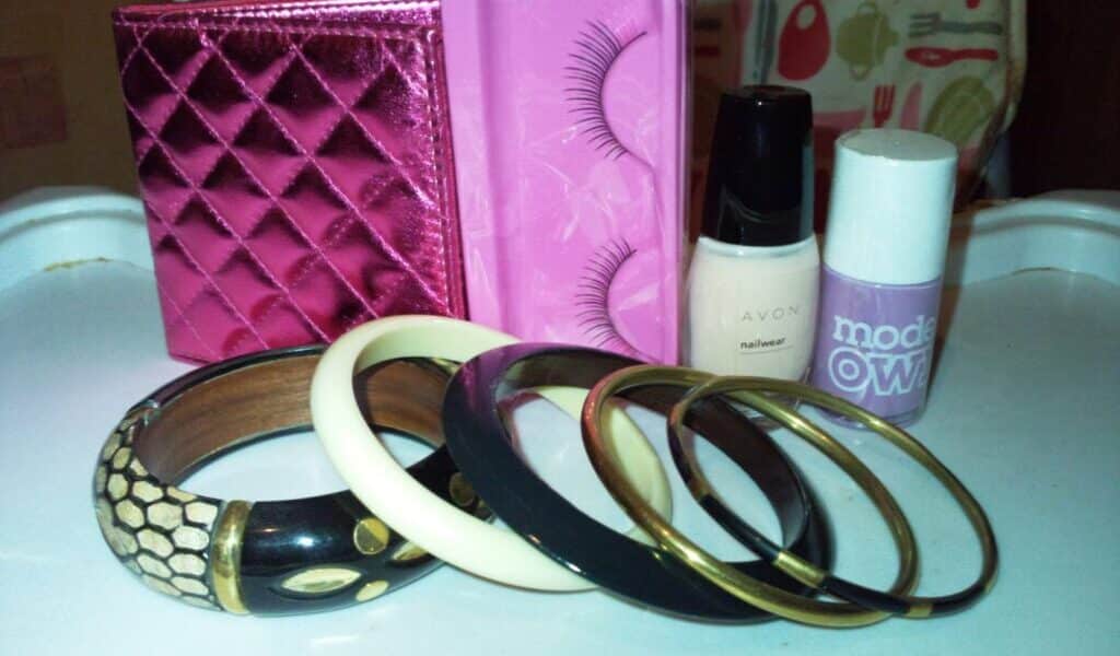 A set of beauty and fashion items on a white surface: a shiny pink quilted purse, a pink box with false eyelashes, Avon nail varnish, Models Own lavender nail varnish, four bangles (wood, bone, and metal), and a wooden bracelet with a snakeskin pattern.