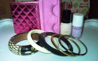 A set of beauty and fashion items on a white surface: a shiny pink quilted purse, a pink box with false eyelashes, Avon nail varnish, Models Own lavender nail varnish, four bangles (wood, bone, and metal), and a wooden bracelet with a snakeskin pattern.