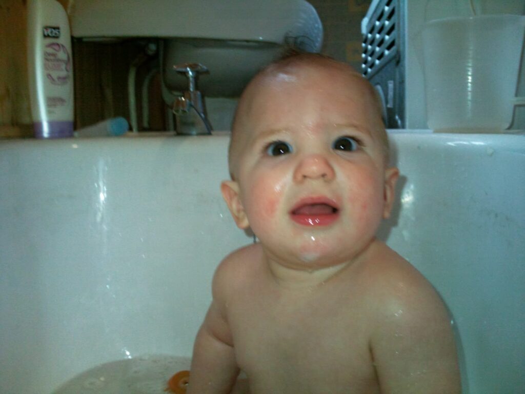 A baby with short hair sits in a white bath, looking slightly upward with a surprised or confused expression. The baby’s mouth is slightly open. In the background are bathroom items, including a bottle and a white basin.