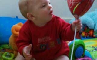 A baby wearing a red shirt sits on a colourful play mat, surrounded by toys. The baby is holding a red heart-shaped balloon with "I Love You" written on it, gazing at the balloon with curiosity. The background shows soft toys and a blue cushion.