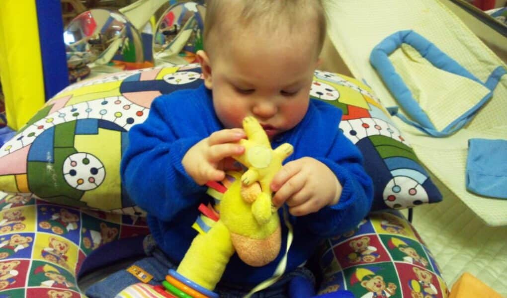A baby in a blue outfit sits on a colourful, patterned cushion holding and examining a soft yellow giraffe toy. The background includes more toys and soft play equipment, creating a playful and cosy atmosphere.