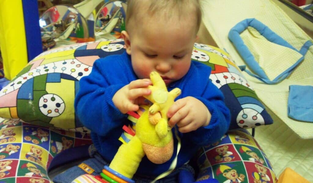 A baby in a blue outfit sits on a colourful, patterned cushion holding and examining a soft yellow giraffe toy. The background includes more toys and soft play equipment, creating a playful and cosy atmosphere.