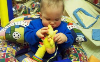 A baby in a blue outfit sits on a colourful, patterned cushion holding and examining a soft yellow giraffe toy. The background includes more toys and soft play equipment, creating a playful and cosy atmosphere.