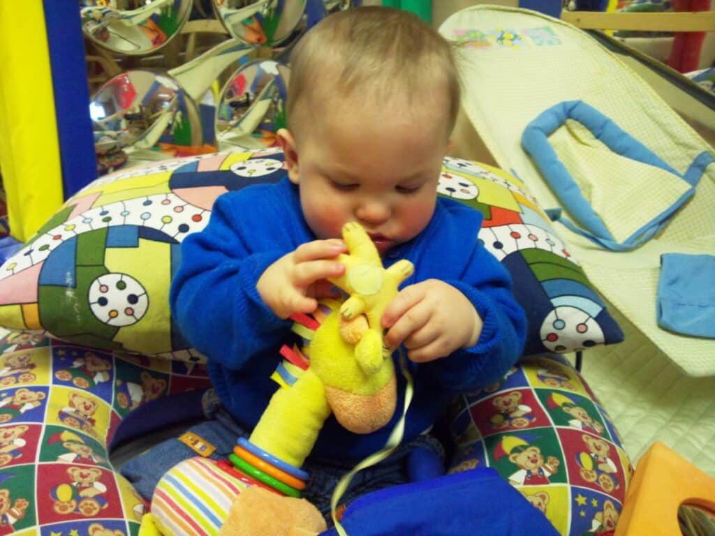 A baby in a blue outfit sits on a colourful cushion, focusing intently on a yellow plush giraffe toy. The room is filled with bright colours, patterns, and soft play equipment, creating a playful, cosy atmosphere.