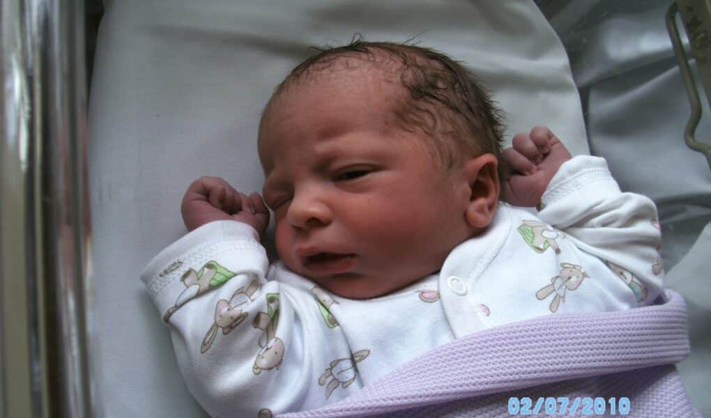 A newborn baby lies in a hospital cot on a white sheet, wearing a white babygrow with bunny patterns and wrapped in a lilac blanket. The baby has a slightly furrowed brow and clenched fists. The date "02/07/2010" is visible in blue text.