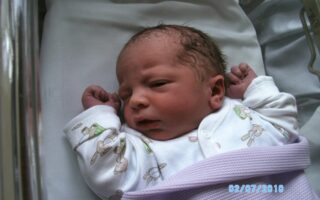 A newborn baby lies in a hospital cot on a white sheet, wearing a white babygrow with bunny patterns and wrapped in a lilac blanket. The baby has a slightly furrowed brow and clenched fists. The date "02/07/2010" is visible in blue text.