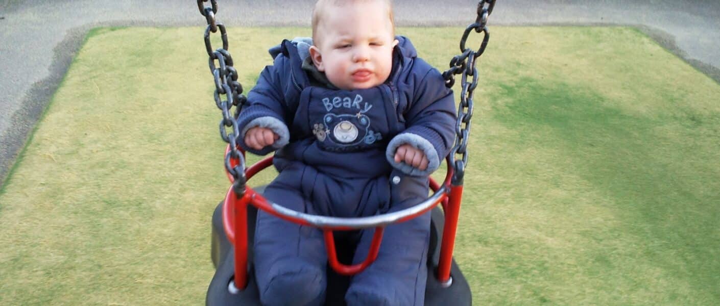 A baby in a padded blue snowsuit sits on a black and red swing at a playground, holding the swing's chains. The baby appears calm, with a slight frown, and there is green artificial grass beneath and a fence in the background.