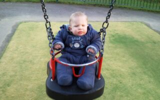 A baby in a padded blue snowsuit sits on a black and red swing at a playground, holding the swing's chains. The baby appears calm, with a slight frown, and there is green artificial grass beneath and a fence in the background.