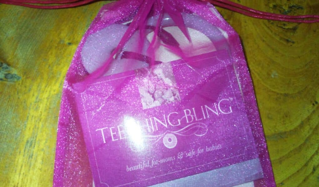 A pink organza gift bag with a drawstring holds a “Teething Bling” product. The packaging reads, “TEETHING BLING, beautiful for mums & safe for babies.” The bag sits on a rustic yellow wooden surface.