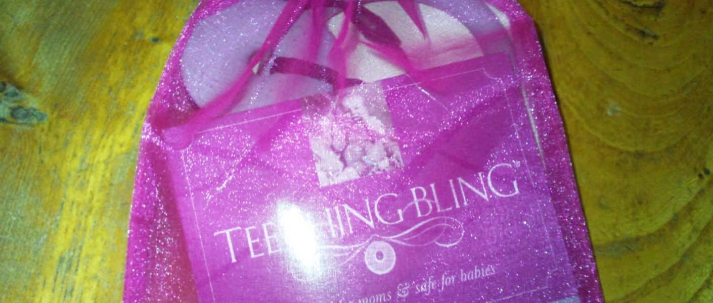 A pink organza gift bag with a drawstring holds a “Teething Bling” product. The packaging reads, “TEETHING BLING, beautiful for mums & safe for babies.” The bag sits on a rustic yellow wooden surface.