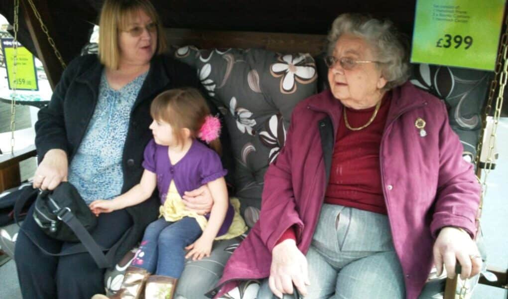 Three generations sit together on a floral cushioned garden swing: an older woman in a maroon coat, a young girl in a purple top, and a woman with blonde hair. Price tags hang nearby, advertising the garden swing for £399.