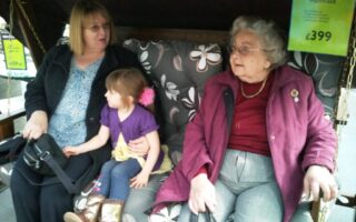 Three generations sit together on a floral cushioned garden swing: an older woman in a maroon coat, a young girl in a purple top, and a woman with blonde hair. Price tags hang nearby, advertising the garden swing for £399.
