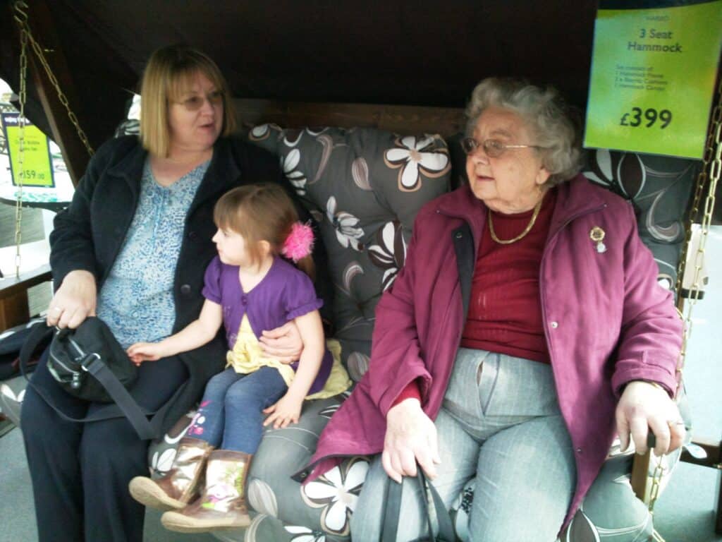 Three people sit on a cushioned outdoor swing: an older woman in a maroon coat, a middle-aged woman in a black coat, and a young girl in a purple shirt and boots. Price signs for hammocks are visible behind them. They appear to be in a shop.