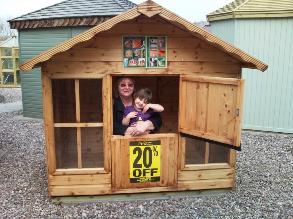 A woman and a young child smile from inside a small wooden playhouse. The playhouse has a window, an open door, and a “20% OFF” sign on the front. Gravel covers the ground, and other shed-like buildings are visible in the background.