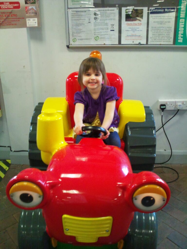 A young girl with brown hair and fringe, wearing a purple top and blue jeans, smiles while sitting on a colourful red and yellow ride-on toy tractor indoors. Notice boards and electrical sockets are visible on the wall behind her.