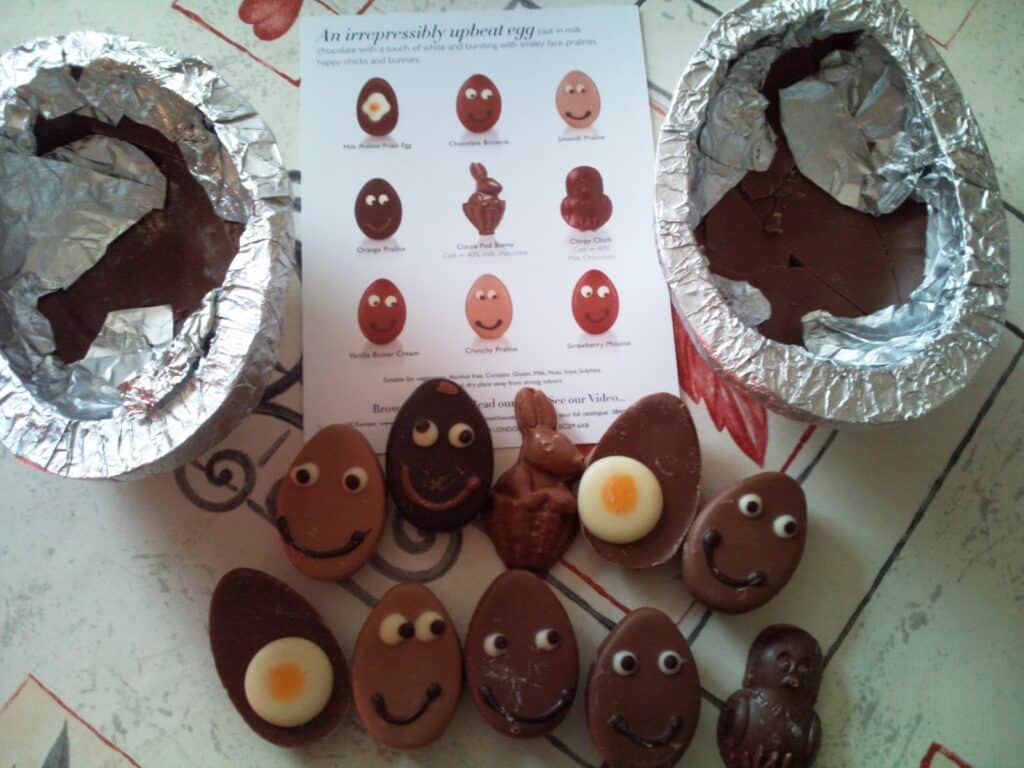 Two large chocolate eggs, broken open with foil lining, surround a group of small chocolate eggs and figures with faces made of white and dark chocolate. A leaflet displaying images and names of the chocolates is placed in the centre.
