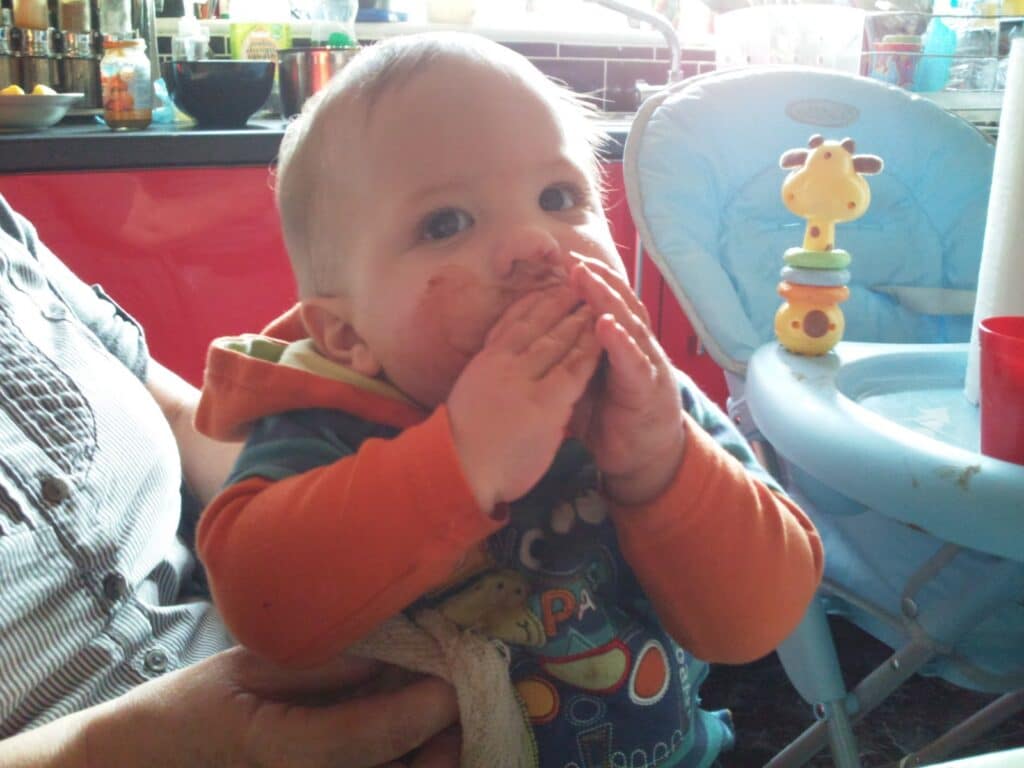 A baby wearing an orange-sleeved shirt sits on an adult’s lap, hands and mouth messy as if just eating. There’s a blue highchair with a giraffe toy beside them, and kitchen items are visible in the background. The baby looks upwards, touching their mouth.
