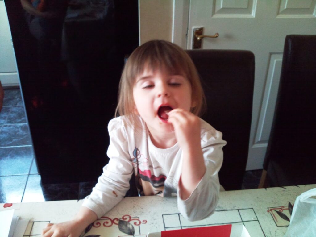 A young girl with brown hair wearing a white long-sleeve shirt sits at a table, eyes half-closed, putting food in her mouth. She appears to be enjoying a snack. The background shows a white door and dark chairs.