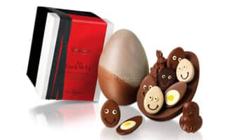 A large, two-toned chocolate egg is cracked open, revealing smaller chocolate pieces inside. The smaller chocolates are egg-shaped or animal-shaped with smiling faces and bright decorations. A red and black gift box stands beside the chocolates.