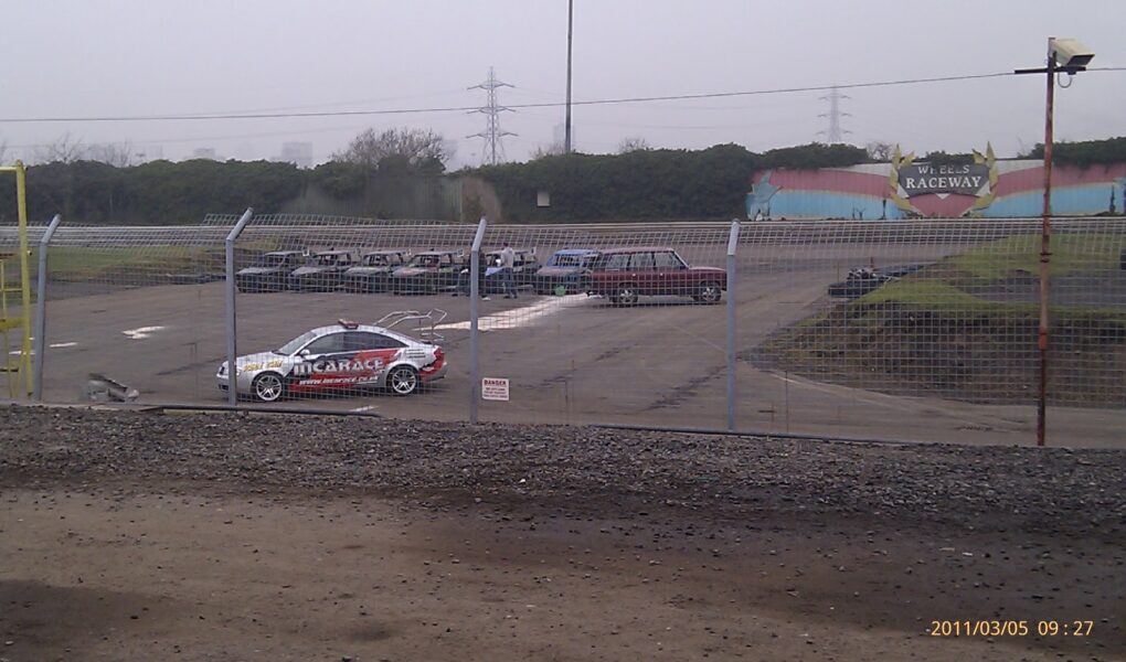 A fenced-off racetrack area with a car labelled “ARC GARAGE” parked on the left, several parked vehicles in the background, and a colourful “ACE CAFE’S RACEWAY” sign on a mural. The sky is overcast and the ground is wet. Timestamp: 2011/03/05 09:27.