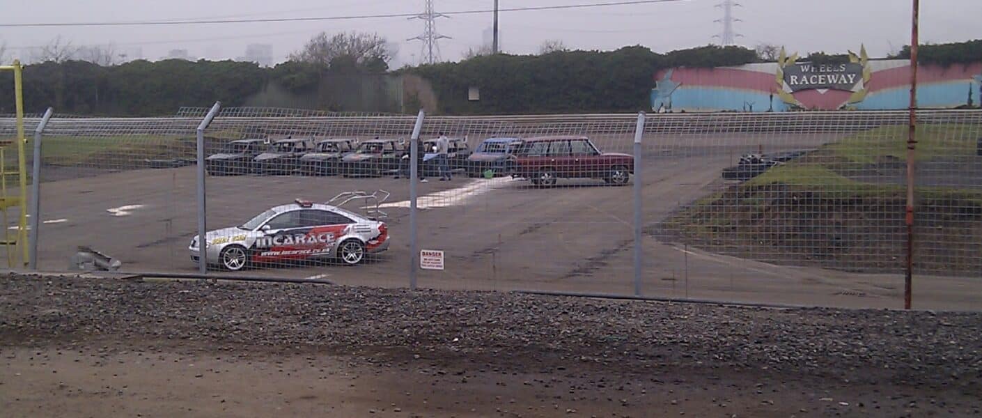 A fenced-off racetrack area with a car labelled “ARC GARAGE” parked on the left, several parked vehicles in the background, and a colourful “ACE CAFE’S RACEWAY” sign on a mural. The sky is overcast and the ground is wet. Timestamp: 2011/03/05 09:27.