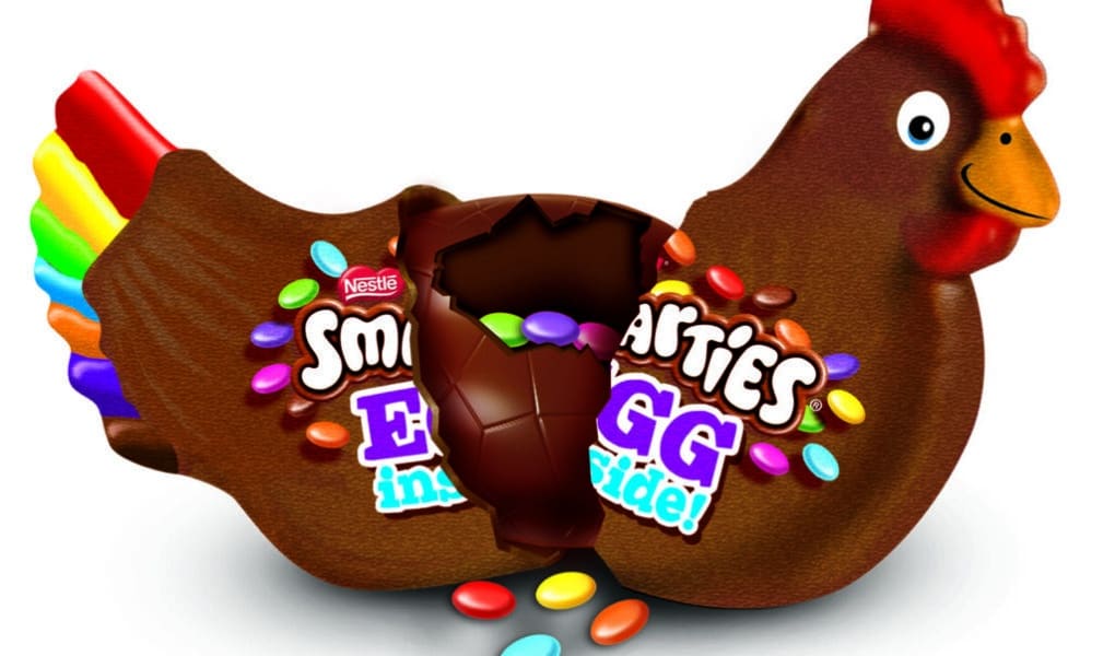 A chocolate hen with a bright red comb and rainbow-coloured tail feathers. Its centre is cracked open, revealing a hollow inside with colourful Smarties spilling out. The label reads “Smarties Egg Inside!” with pieces of chocolate and sweets scattered around.
