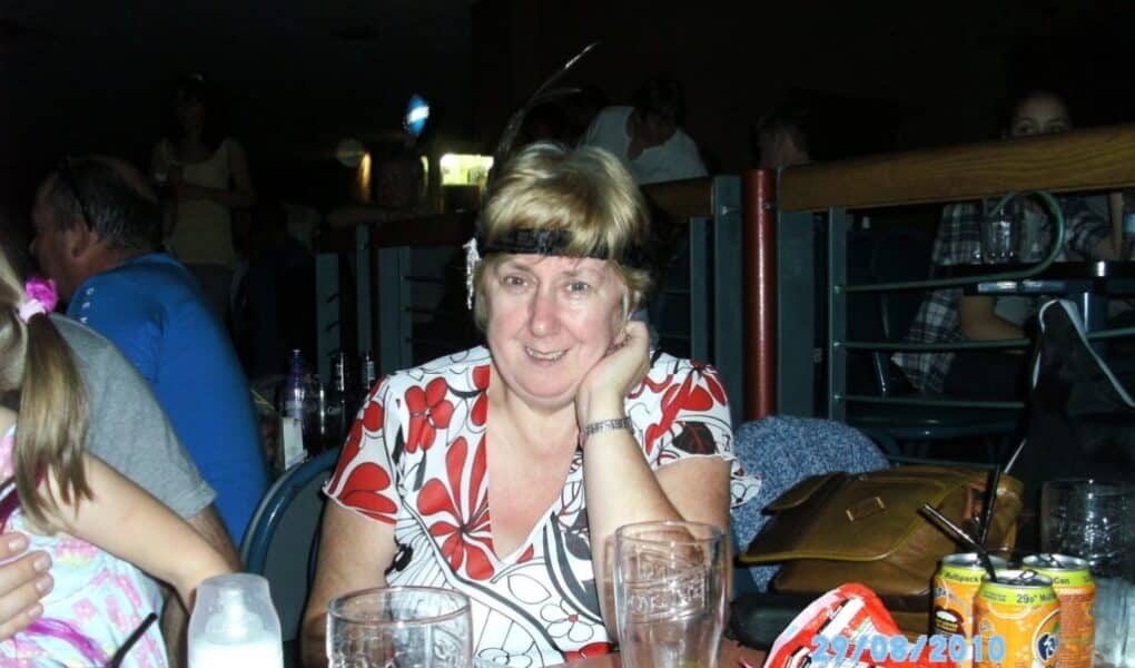 A woman with short blonde hair, wearing a white and red patterned top, sits at a table with empty glasses, fizzy drink cans, and plastic containers. She is smiling and resting her face on her hand. The setting appears to be a dimly lit indoor venue.