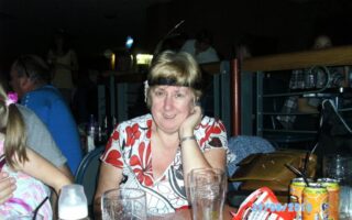 A woman with short blonde hair, wearing a white and red patterned top, sits at a table with empty glasses, fizzy drink cans, and plastic containers. She is smiling and resting her face on her hand. The setting appears to be a dimly lit indoor venue.