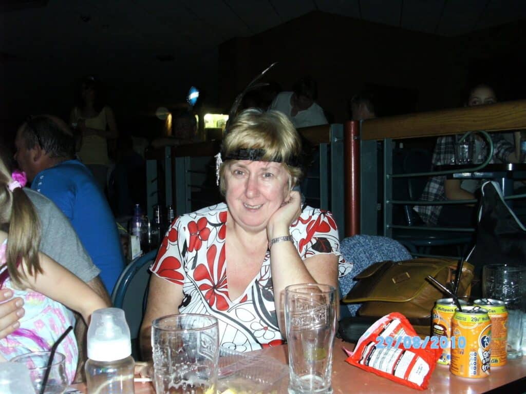 A smiling woman with short blonde hair and a floral top sits at a table with empty glasses, drinks, and snacks. She rests her head on her hand. Other people are in the background, and the date "27/08/2010" is visible on the lower right-hand side.