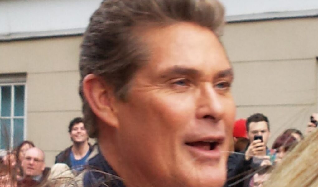 A man with short, styled brown hair is smiling amid a crowd outdoors. Several people in the background are taking photos with their mobiles. The setting includes a beige building with a window and white trim. The image is slightly blurred.