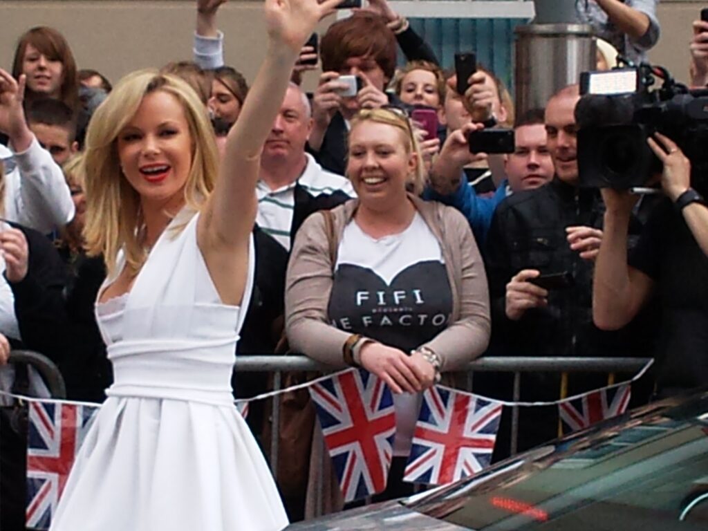 A woman in a white dress waves and smiles at a crowd. People behind a barrier hold phones and cameras, taking photos. British flags hang on the railing, and a camera operator films the scene. The atmosphere is lively and celebratory.
