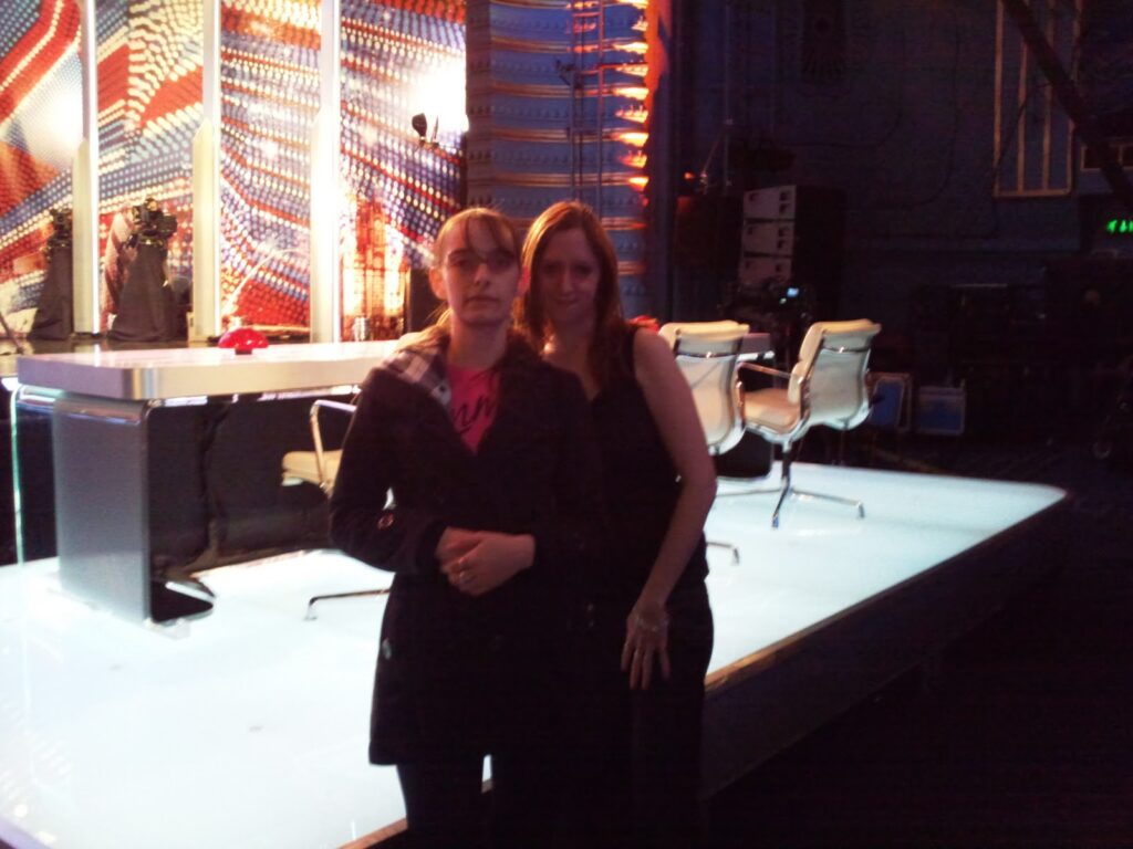 Two women stand together in front of a brightly lit judges’ table on a TV talent show set. The background features colourful, illuminated patterns and empty seats behind the table. The setting appears backstage or during a break in filming.