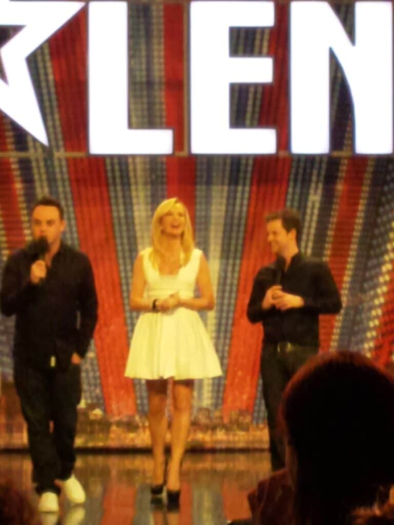 Three people stand onstage in front of a Britain’s Got Talent backdrop. The woman in the centre wears a short white dress and black heels. The two men on either side wear black outfits and hold microphones. The background features large letters and a red, white, and blue design.